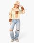 A woman modeling a cream and brown patchwork knit cardigan with a beige fringed bucket hat and distressed light-wash jeans, standing against a plain white studio background.