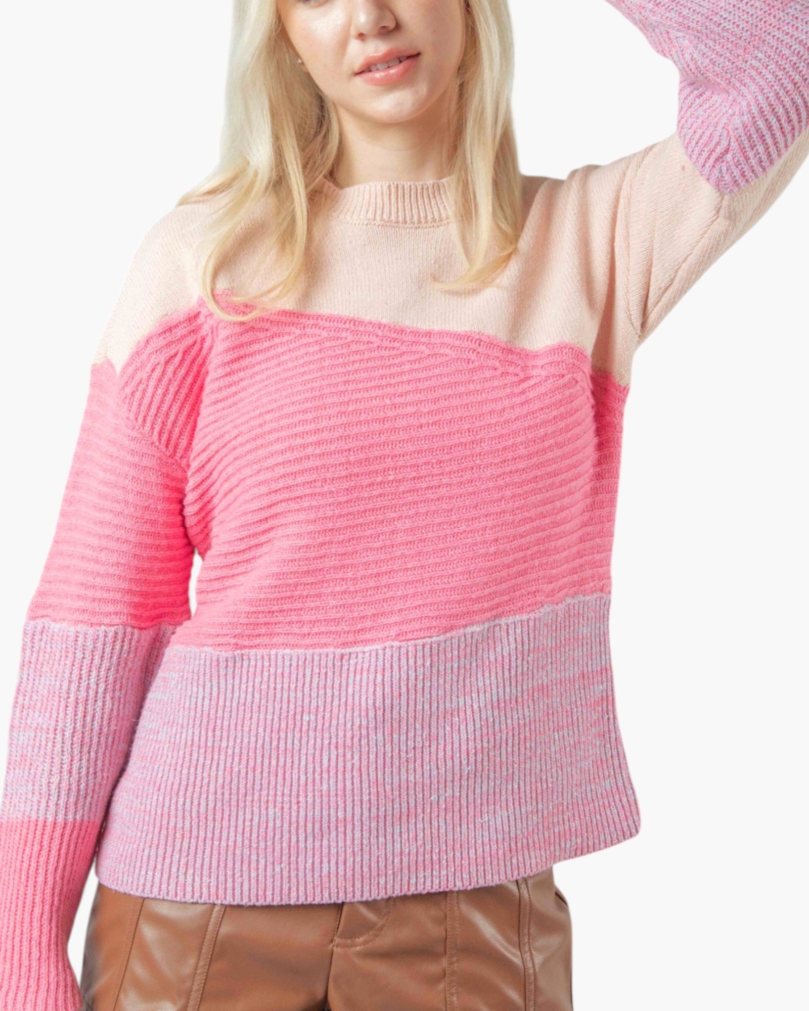 A woman wearing a color-blocked ribbed knit sweater with a light pink upper section, a brighter pink middle section, and a lavender-pink lower section and sleeves, paired with brown faux-leather pants against a white background.