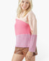 A woman wearing a color-block knit sweater with a light pink upper section, a brighter pink ribbed middle section, and a lavender-pink ribbed lower section, paired with brown faux-leather shorts against a white studio background.