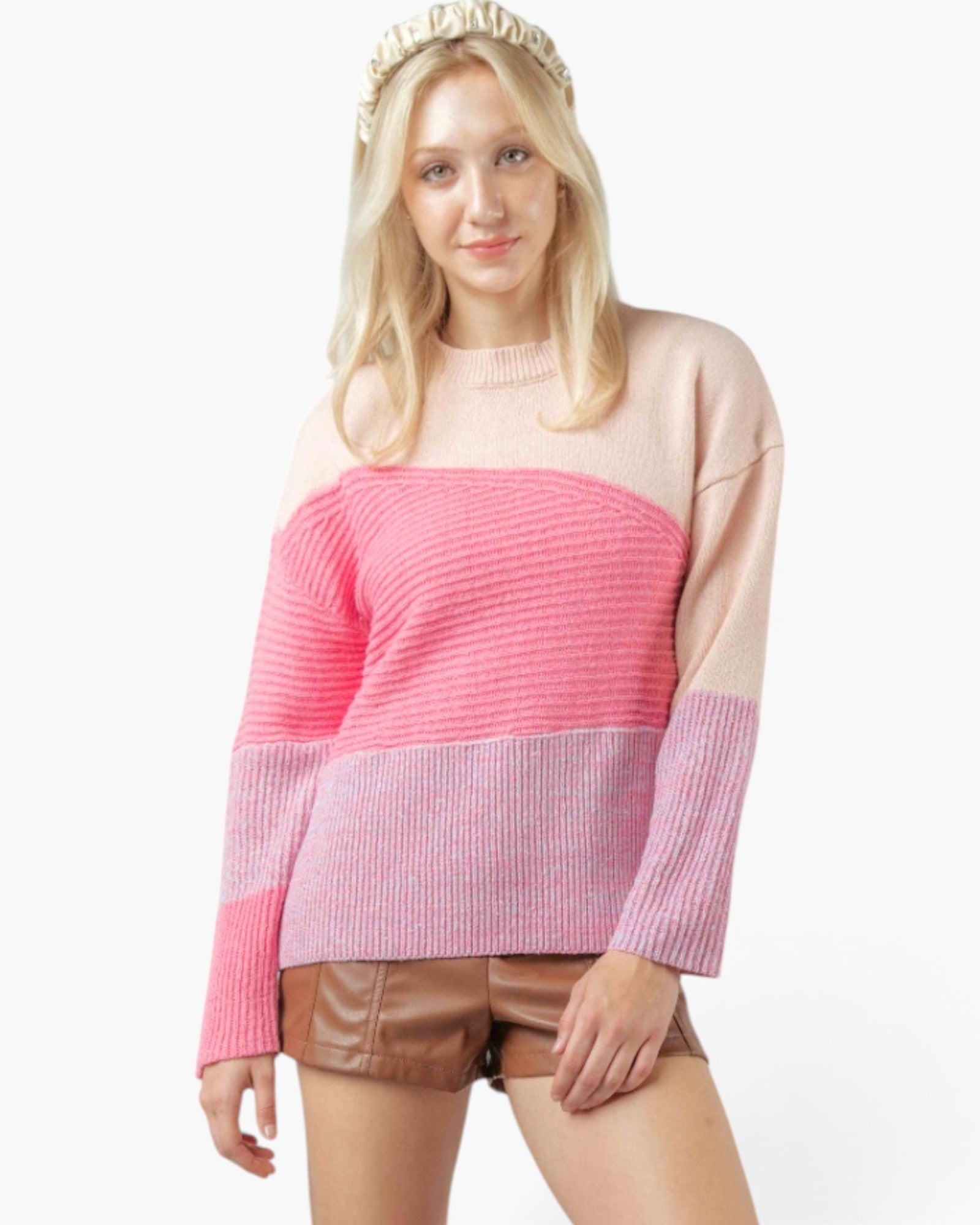 A woman wearing a color-block knit sweater with a light pink upper section, a bright pink ribbed chest band, and pink-and-white striped lower sleeves, paired with brown shorts and a gold beaded headband against a white studio background.
