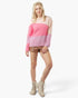 A woman modeling a color-blocked knit sweater with a beige top section, a wide pink middle band, and a pink-and-white gingham lower section, paired with brown shorts, beige lace-up ankle boots, and a beige headband against a light gray studio background.