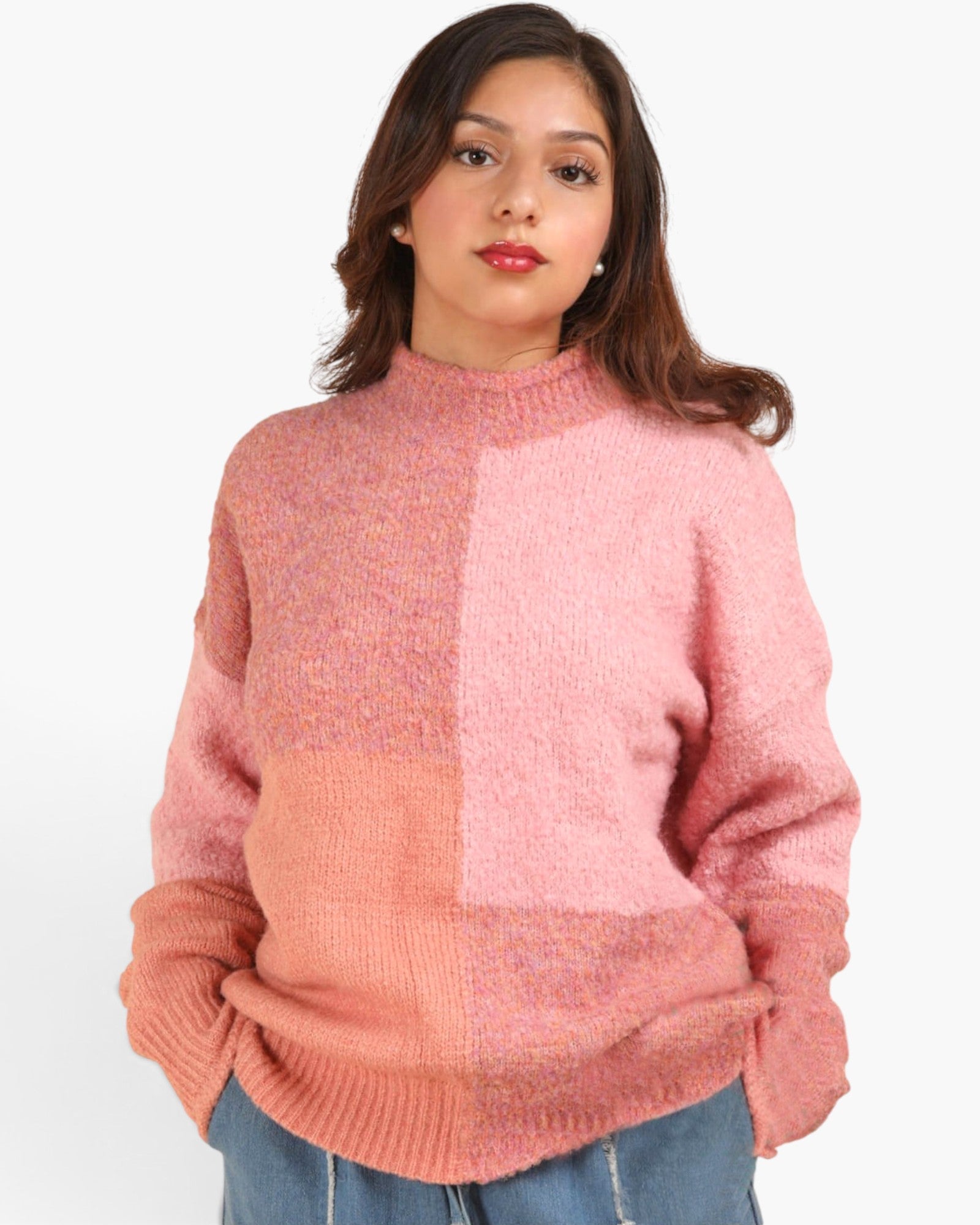A woman wearing a chunky knit mock-neck sweater with a color-blocked design in shades of dusty pink and peach, paired with blue jeans, against a plain white background.