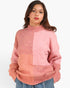 A woman wearing a chunky knit mock-neck sweater with a color-blocked design in shades of dusty pink and peach, paired with blue jeans, against a plain white background.