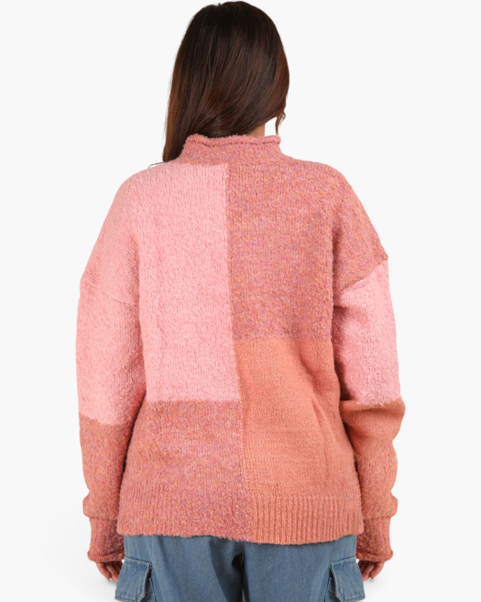 A woman viewed from behind wearing a chunky knit patchwork sweater in shades of pink and peach, paired with blue jeans, against a plain white background.