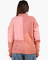 A woman viewed from behind wearing a chunky knit patchwork sweater in shades of pink and peach, paired with blue jeans, against a plain white background.
