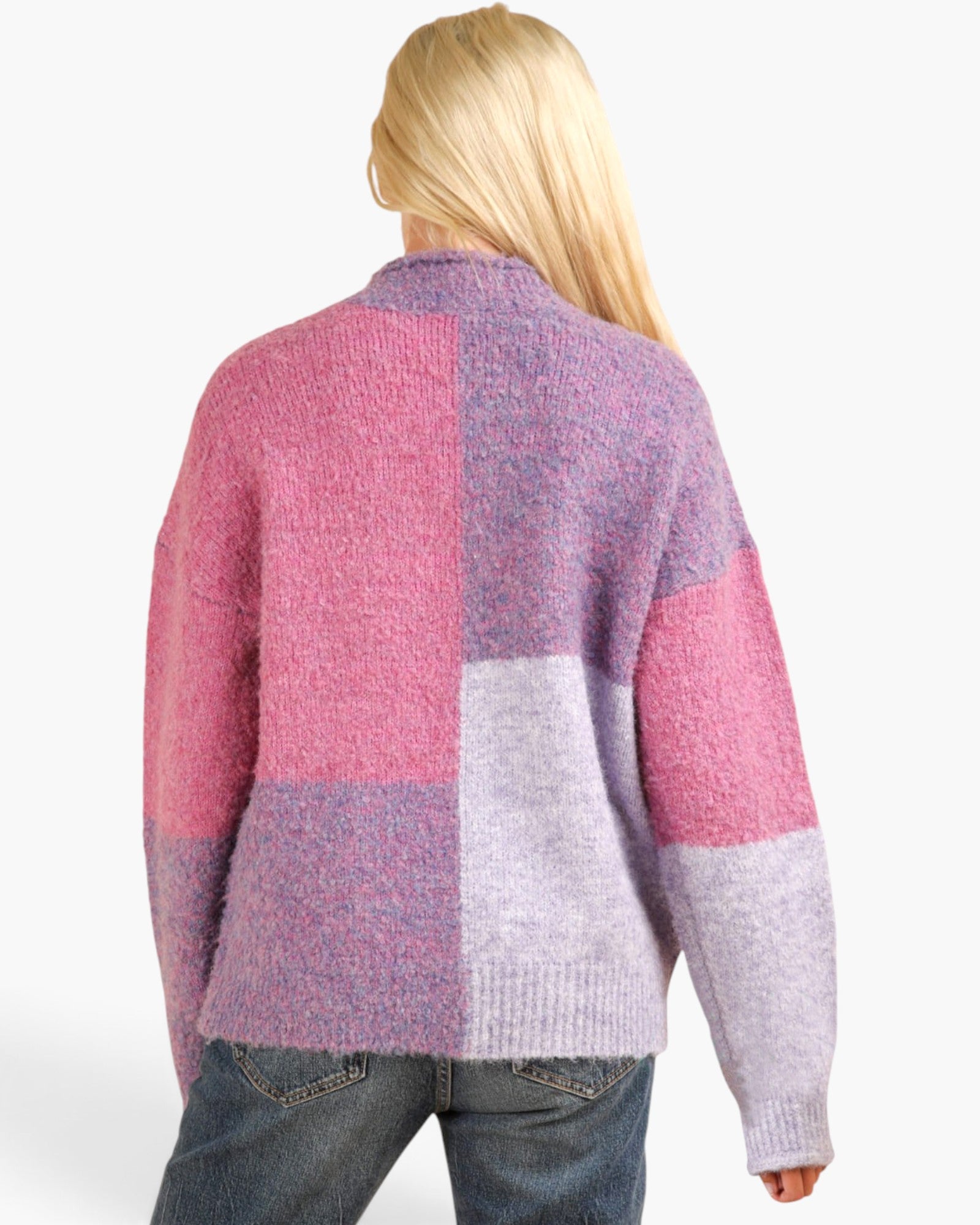 A woman viewed from the back wearing a fuzzy patchwork knit cardigan with pink, lavender, and light gray blocks, paired with blue jeans against a plain white background.