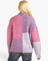A woman viewed from the back wearing a fuzzy patchwork knit cardigan with pink, lavender, and light gray blocks, paired with blue jeans against a plain white background.