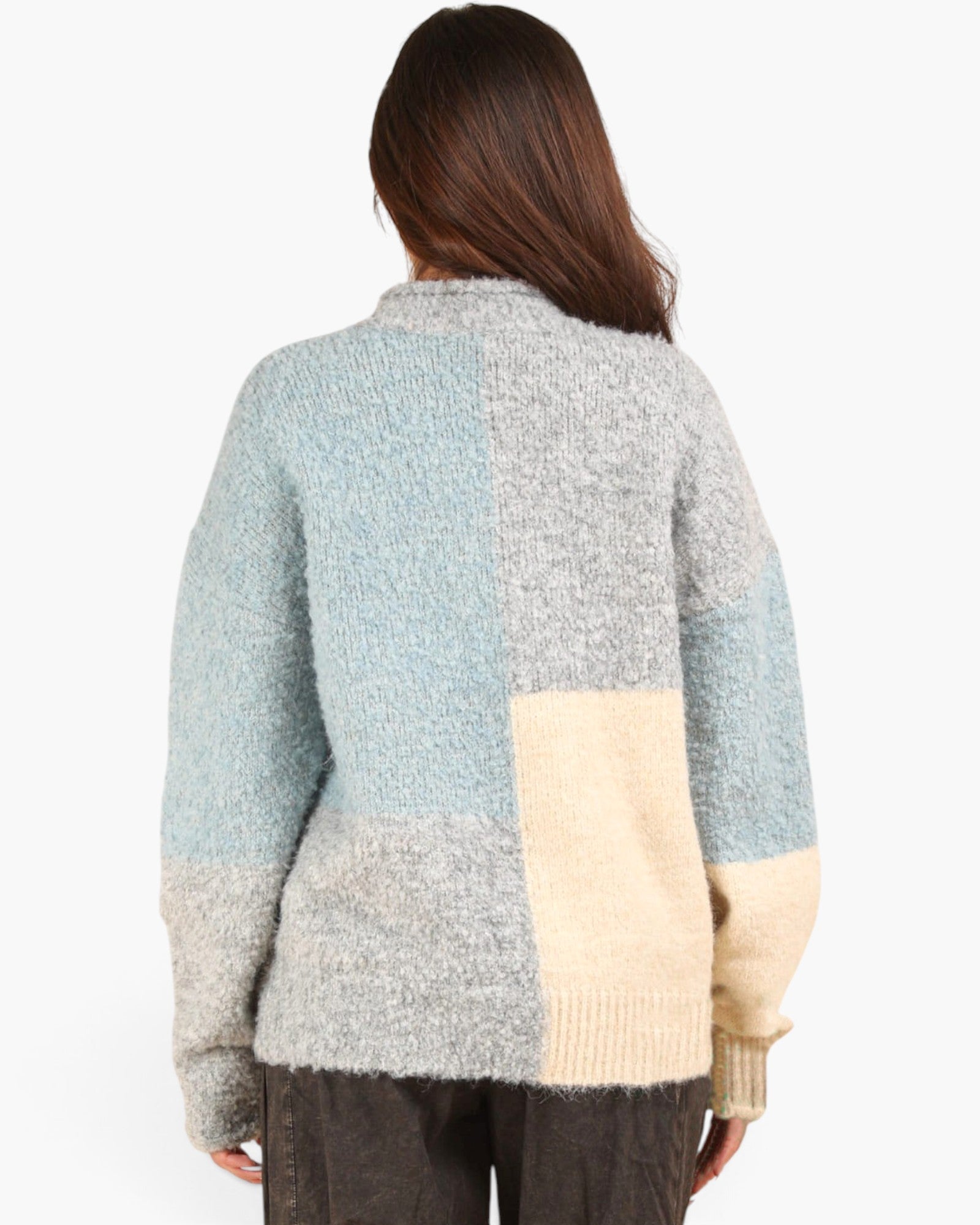 A woman viewed from behind wearing a fuzzy knit patchwork cardigan with blocks of light blue, heather gray, and cream, paired with dark brown pants against a plain white background.