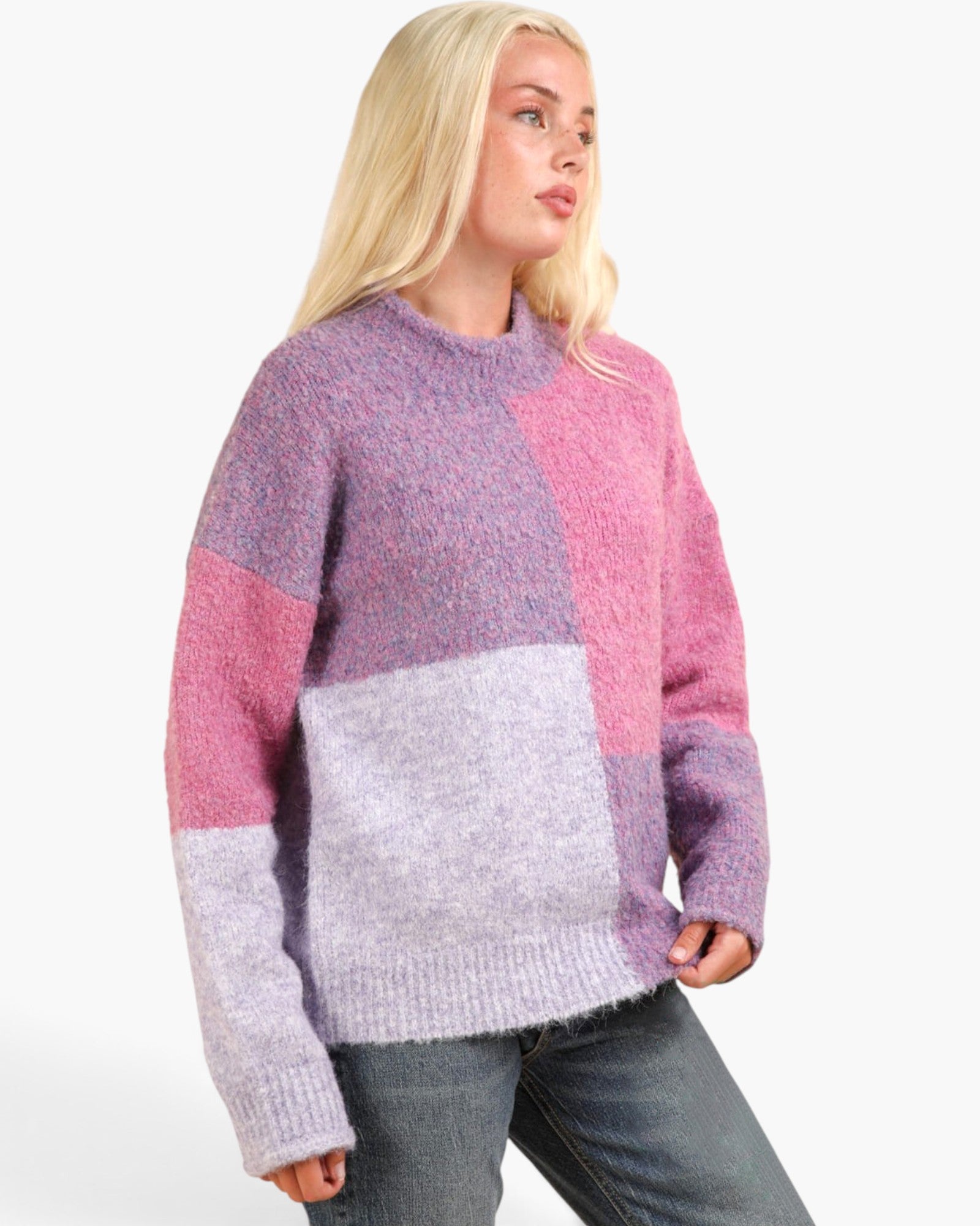 A woman wearing a chunky knit colorblock sweater with pink, purple, and light lavender panels, paired with blue jeans, against a plain white background.