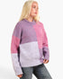A woman wearing a chunky knit colorblock sweater with pink, purple, and light lavender panels, paired with blue jeans, against a plain white background.