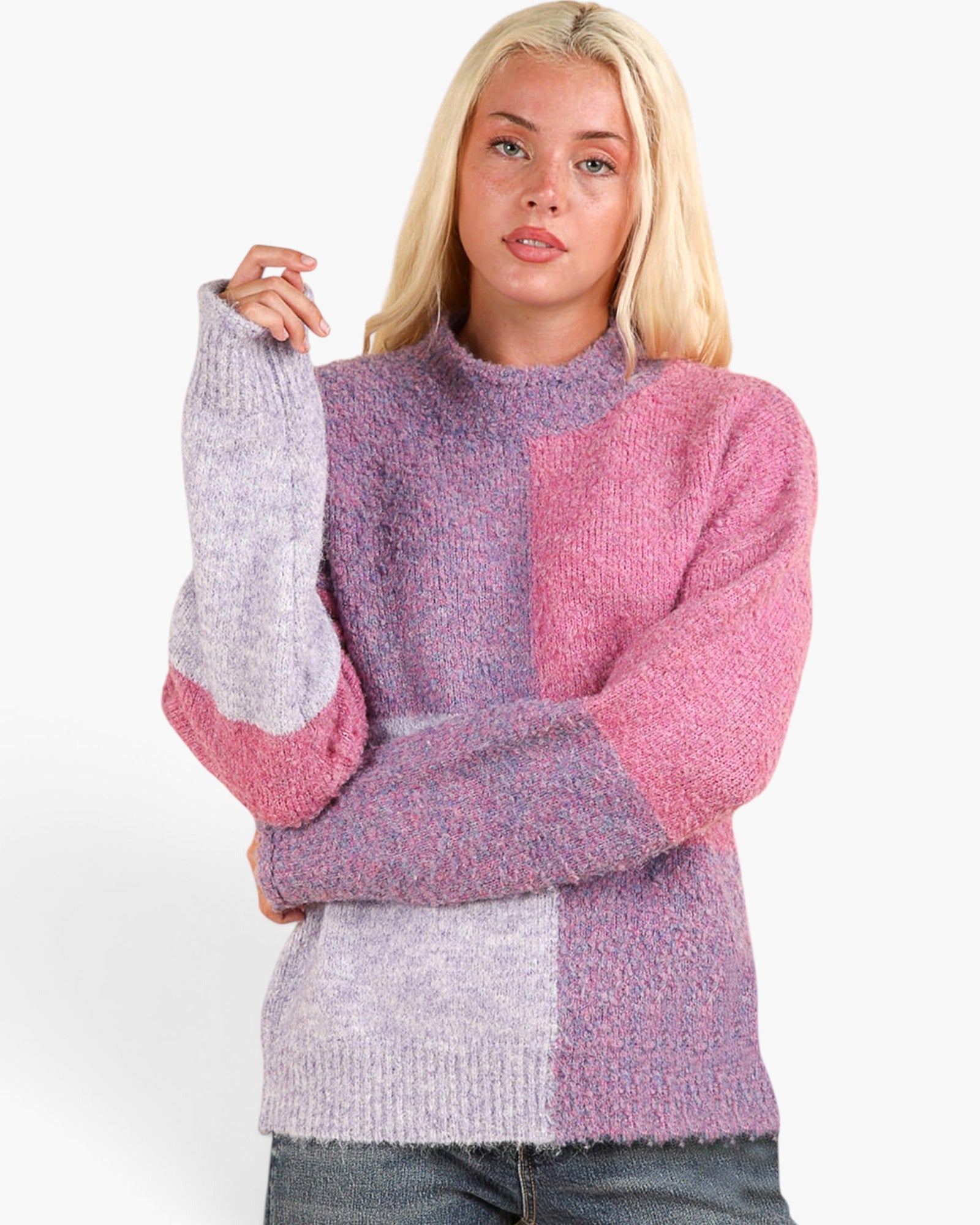 A blonde woman wearing a chunky knit color-block sweater with lavender, pink, and purple panels, paired with dark wash jeans, against a plain white studio background.