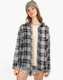 A woman wearing a black and white plaid button-up shirt jacket with distressed details, layered over a cream ribbed top and paired with denim shorts and a green beanie, against a plain white background.