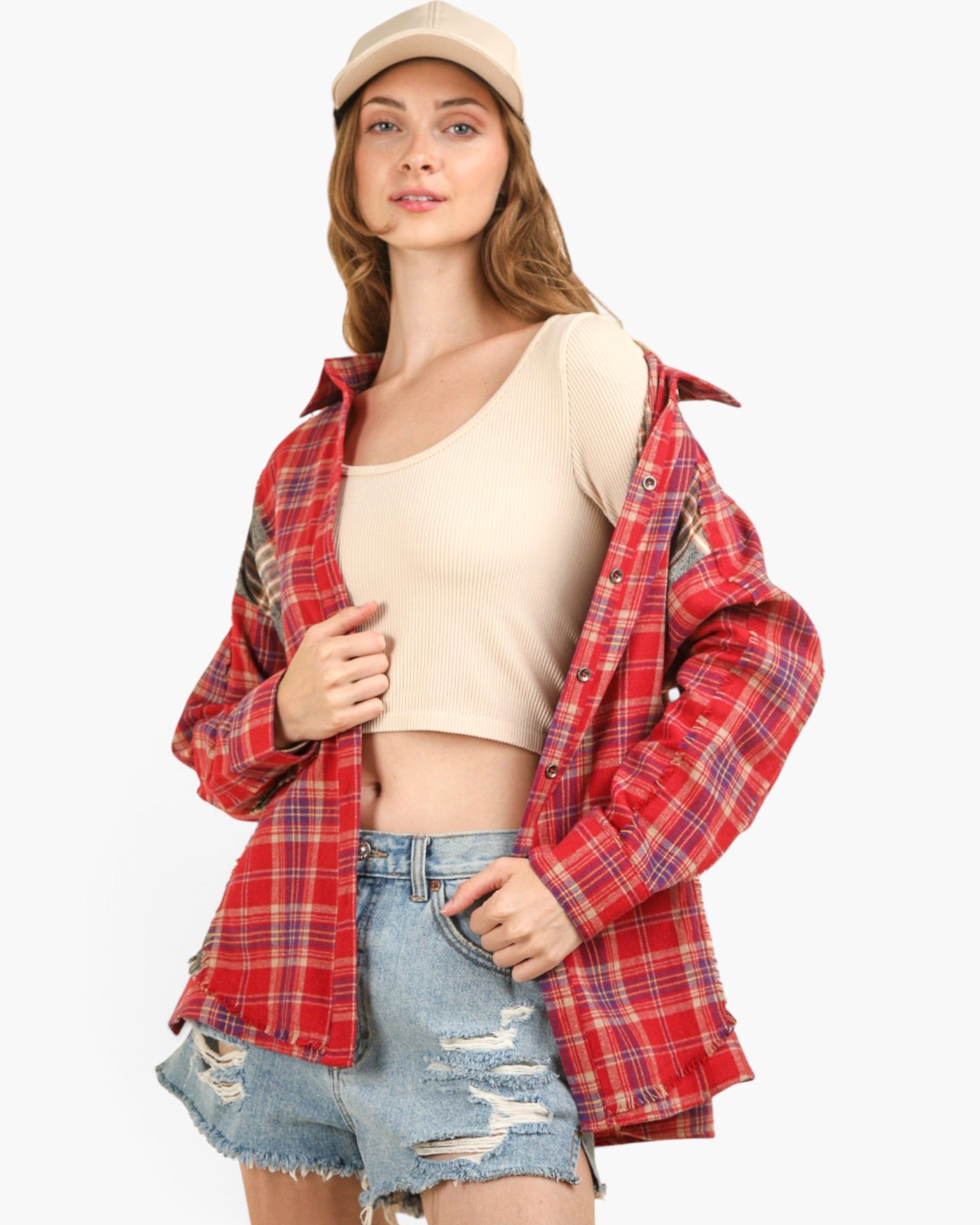 A woman modeling a red and blue plaid flannel shirt jacket worn open over a beige crop top and distressed denim shorts, with a beige cap, against a plain light gray background.