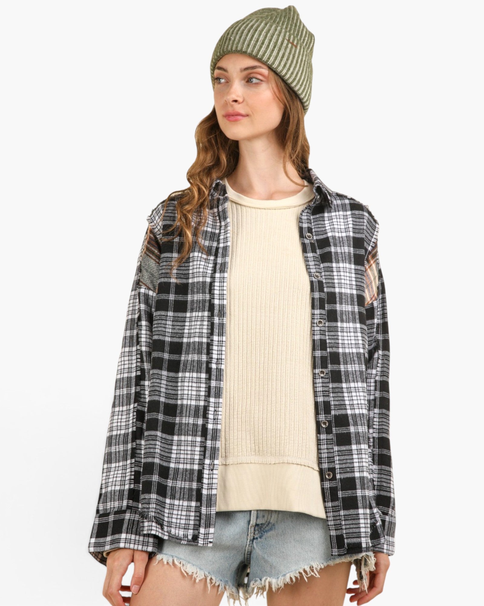 A woman wearing a black and white plaid distressed flannel shirt over a cream ribbed sweater, paired with denim shorts and a green striped beanie, against a plain white background.
