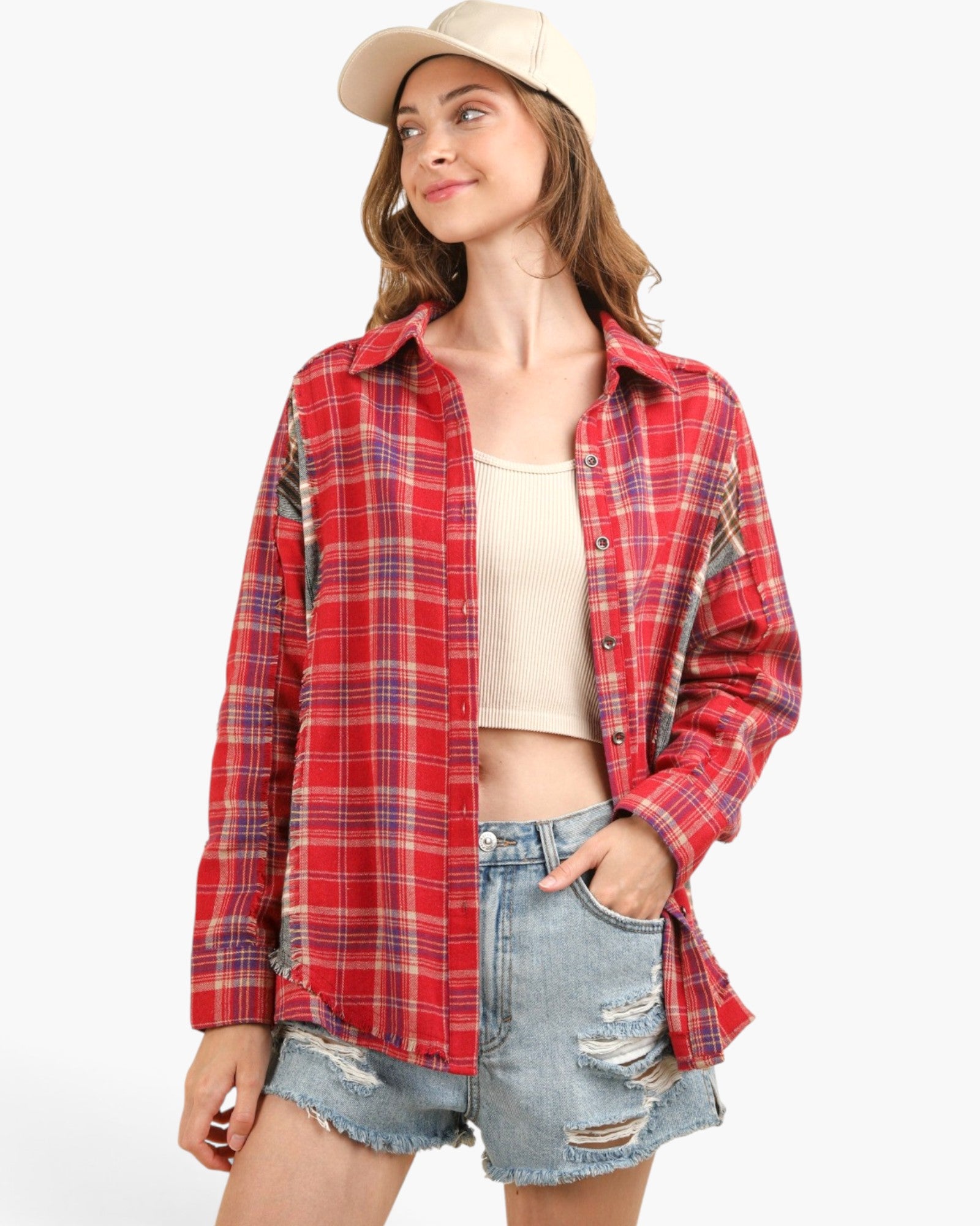 A woman wearing a red and blue plaid button-up flannel shirt over a beige ribbed crop top, paired with distressed denim shorts and a cream baseball cap, set against a plain white studio background.