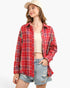 A woman wearing a red and blue plaid button-up flannel shirt over a beige ribbed crop top, paired with distressed denim shorts and a cream baseball cap, set against a plain white studio background.