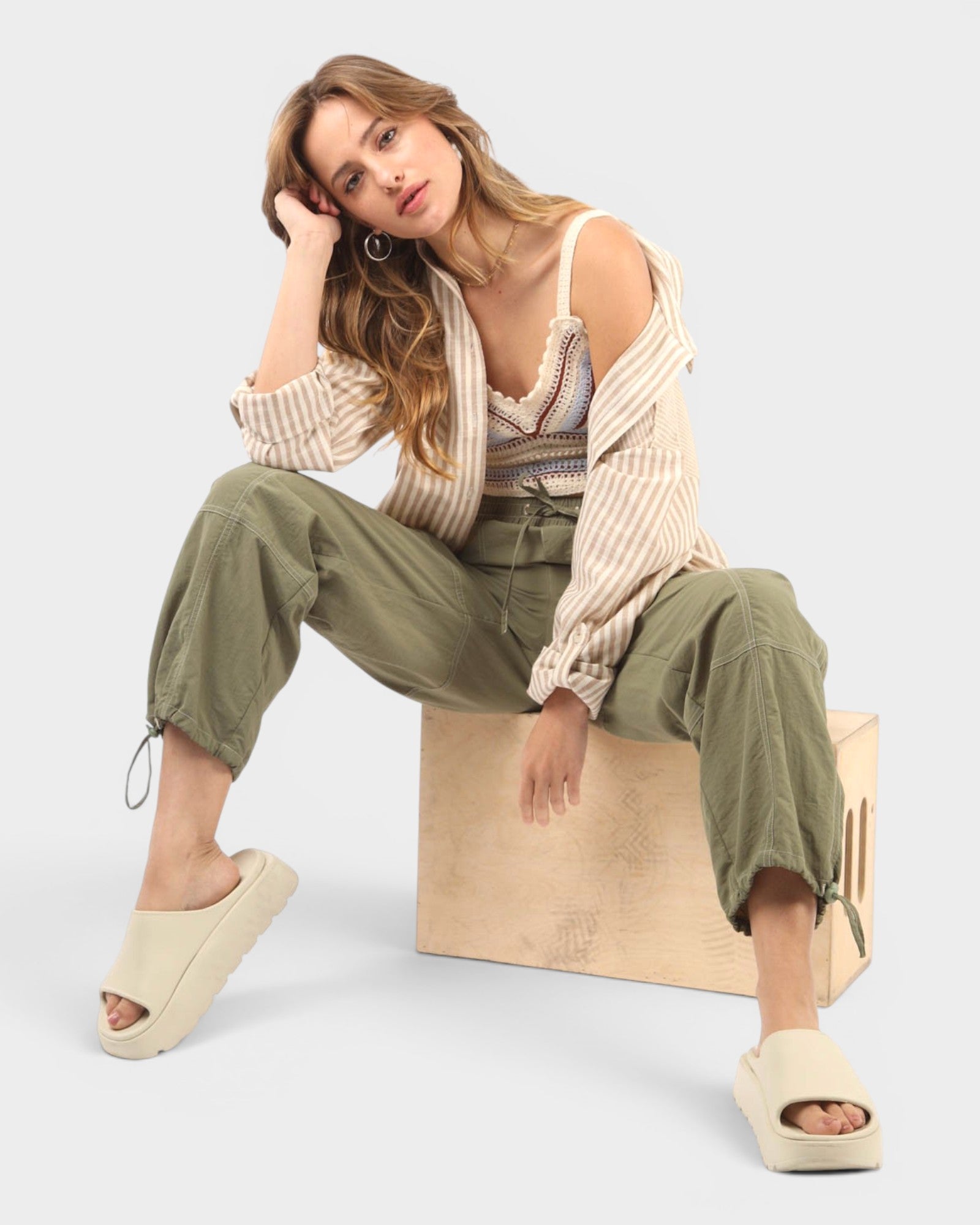 Woman sitting on a box wearing a beige top, green pants, and white sandals on a light gray background