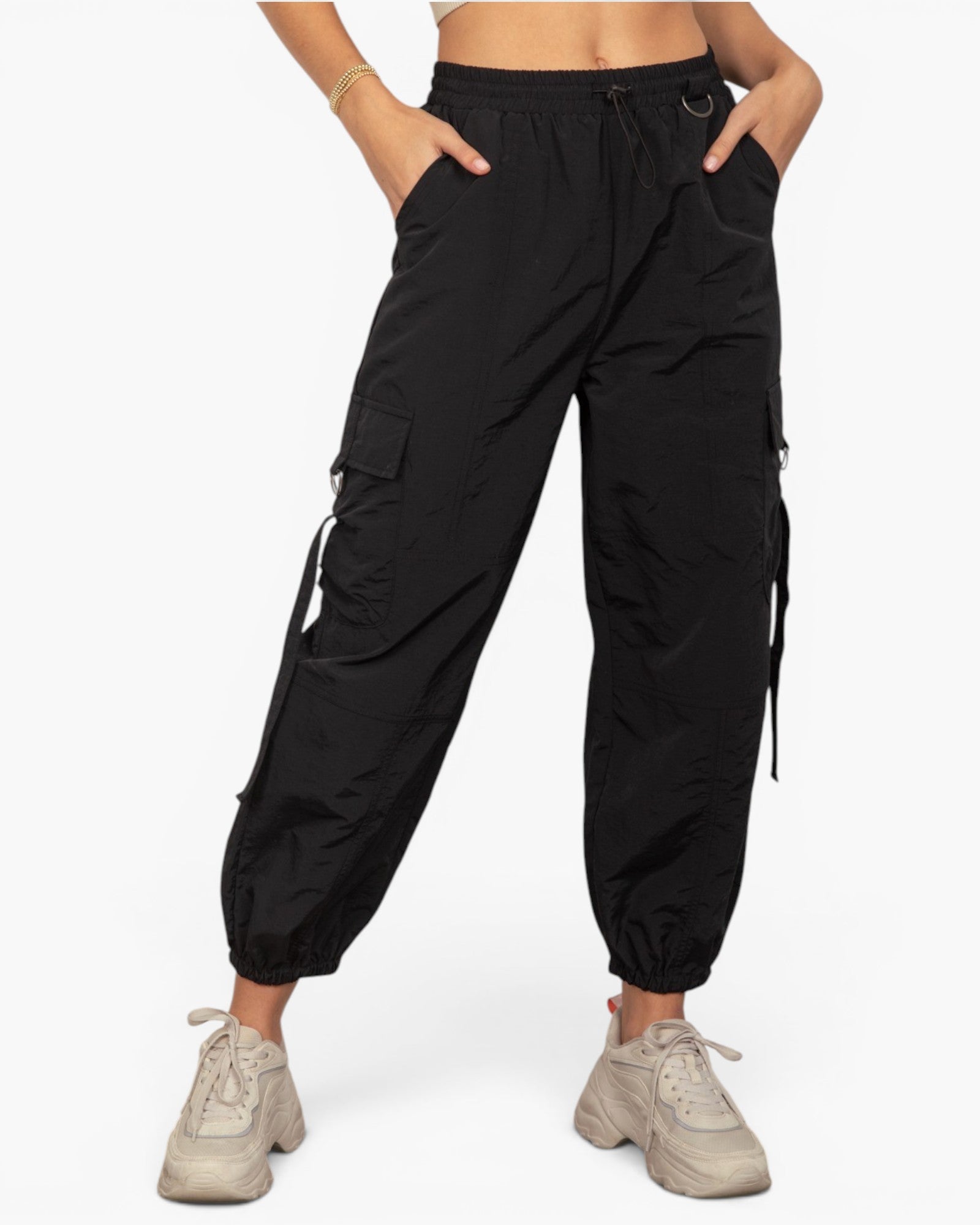 A person wearing black cargo jogger pants with side flap pockets and drawstring details, paired with light beige sneakers, shown against a plain light gray studio background.