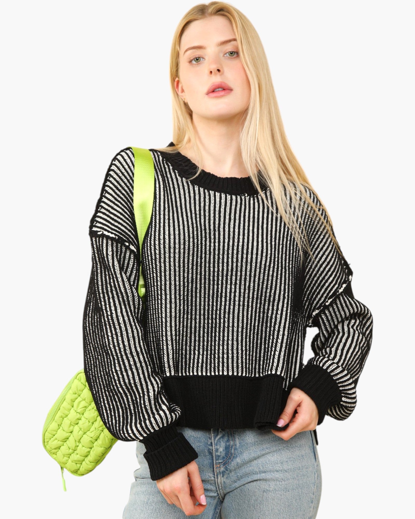 A blonde woman wearing a black and white vertically striped knit sweater with black ribbed trim, paired with light blue jeans and carrying a bright lime green quilted shoulder bag, set against a plain white background.