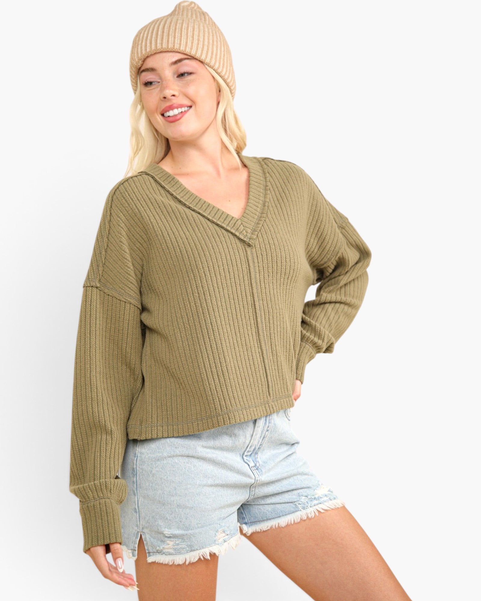 A woman wearing a ribbed knit V-neck sweater in olive green, paired with light-wash denim shorts and a beige beanie, against a plain white background.