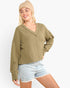 A woman wearing a ribbed knit V-neck sweater in olive green, paired with light-wash denim shorts and a beige beanie, against a plain white background.