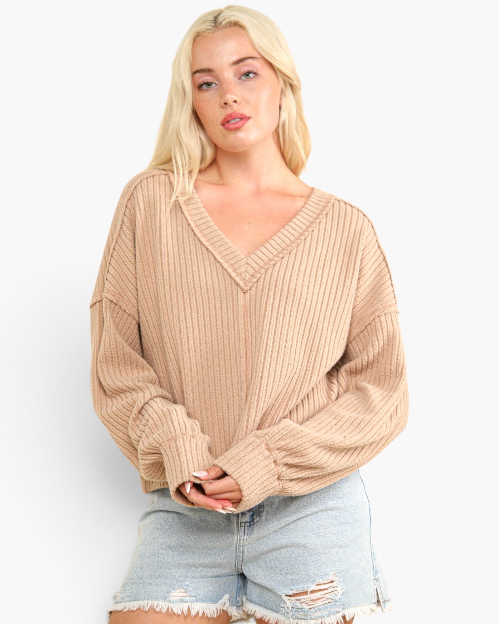 A blonde woman wearing a beige ribbed knit V-neck sweater with balloon sleeves and light-wash distressed denim shorts, standing against a plain white studio background.
