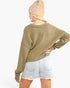 A woman shown from the back wearing an olive green ribbed knit sweater, a beige beanie, and light-wash denim shorts against a plain white background.