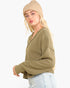 A woman wearing a ribbed knit long-sleeve sweater in olive green, a beige beanie, and light blue jeans, photographed against a plain light gray studio background.