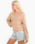 A blonde model wearing a beige ribbed knit V-neck sweater with long sleeves and light-wash distressed denim shorts, photographed against a plain white background.