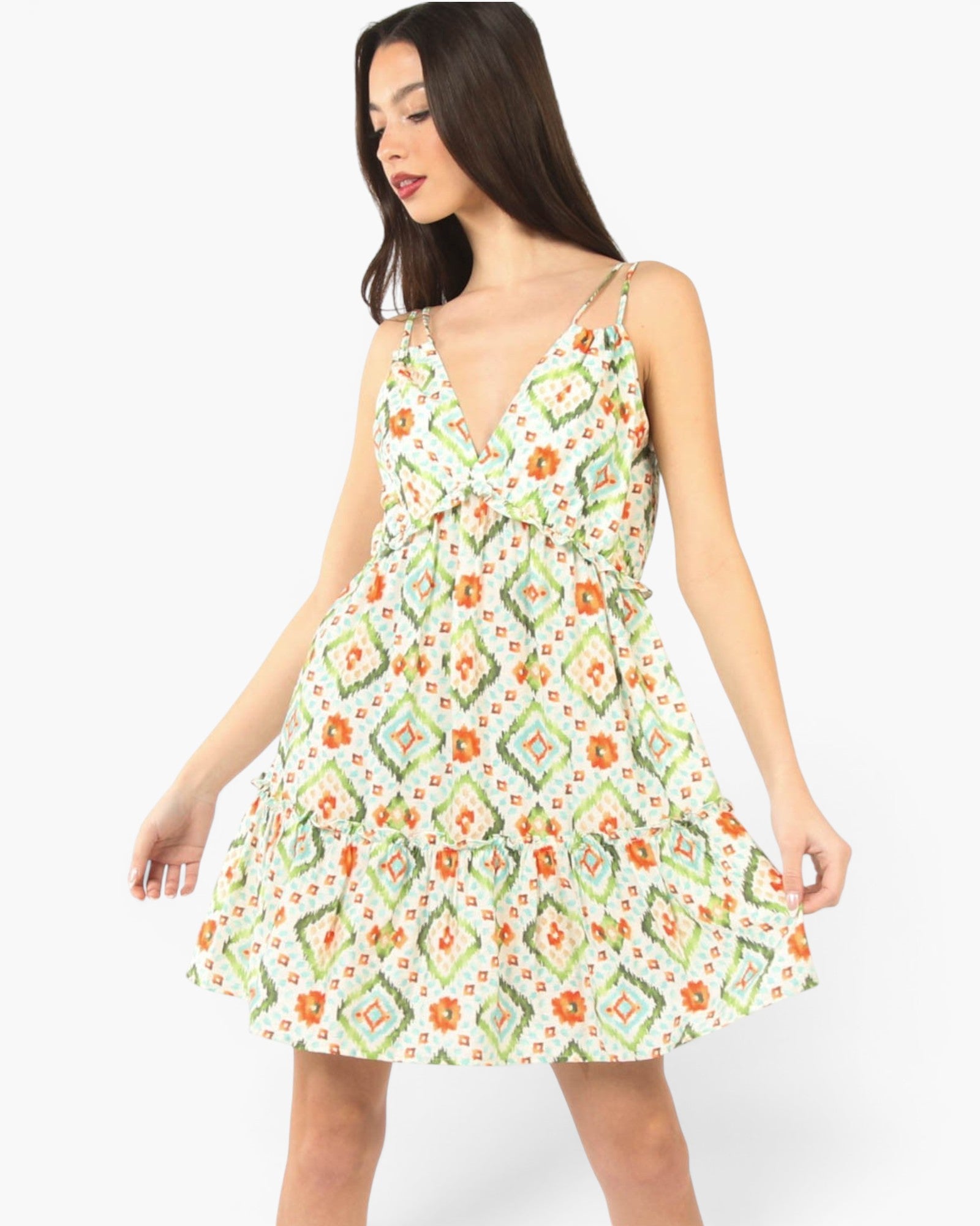 A woman modeling a sleeveless V-neck mini dress with thin straps and a flared skirt, featuring an all-over geometric and floral print in orange, green, and red on a white background, set against a plain light gray studio backdrop.