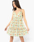 A woman modeling a sleeveless V-neck mini dress with thin straps and a flared skirt, featuring an all-over geometric and floral print in orange, green, and red on a white background, set against a plain light gray studio backdrop.