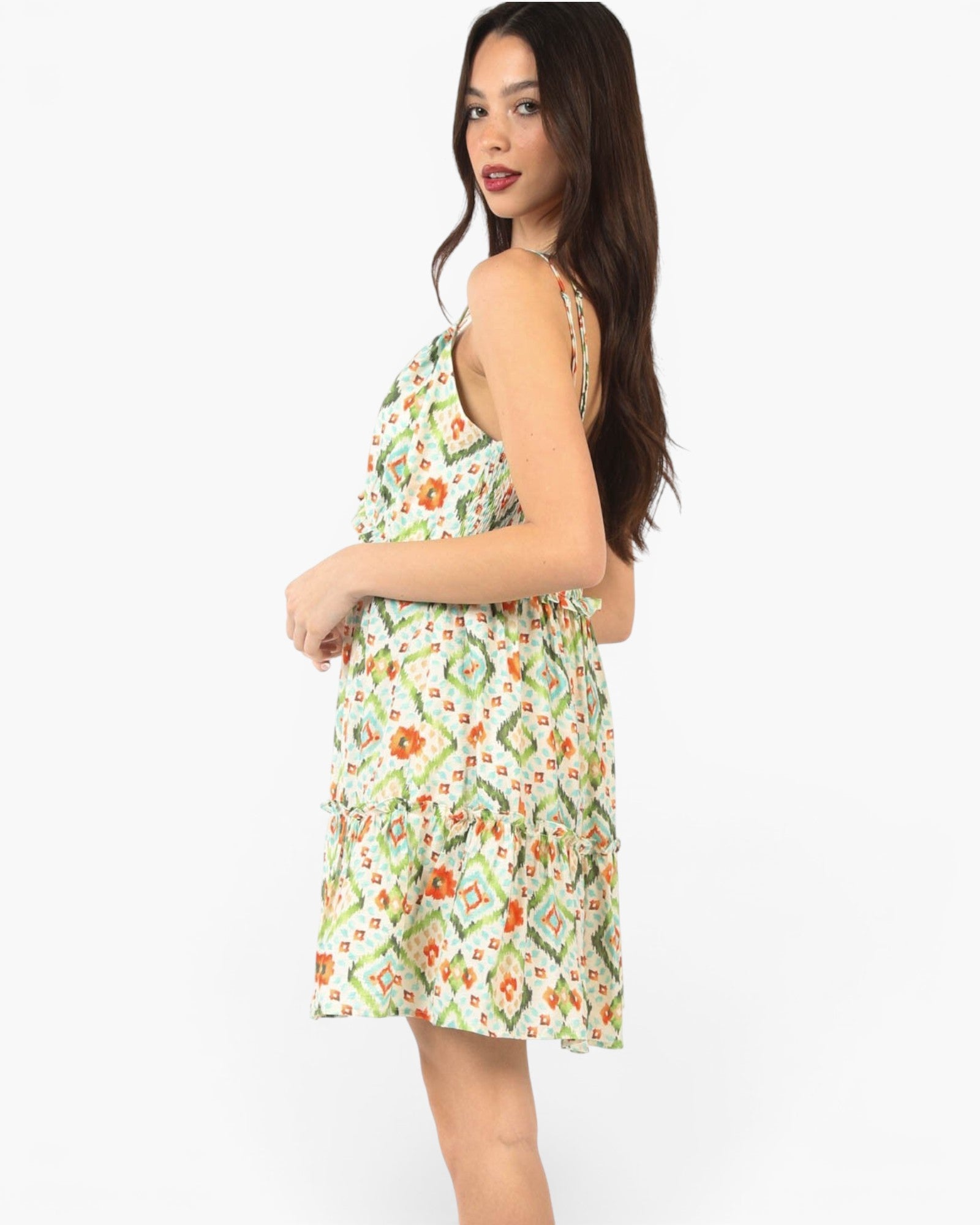 A woman modeling a sleeveless tiered mini dress with a green, orange, and white geometric floral pattern against a plain white background.