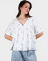Woman wearing a white blouse with blue floral patterns and light blue jeans on a plain background