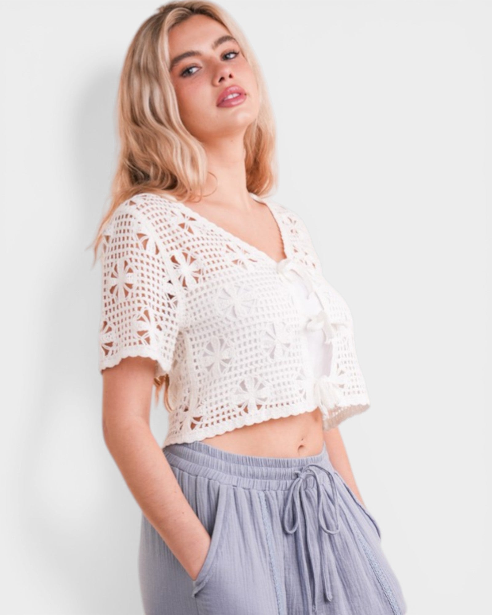 Woman wearing a white crochet top and gray pants against a light background