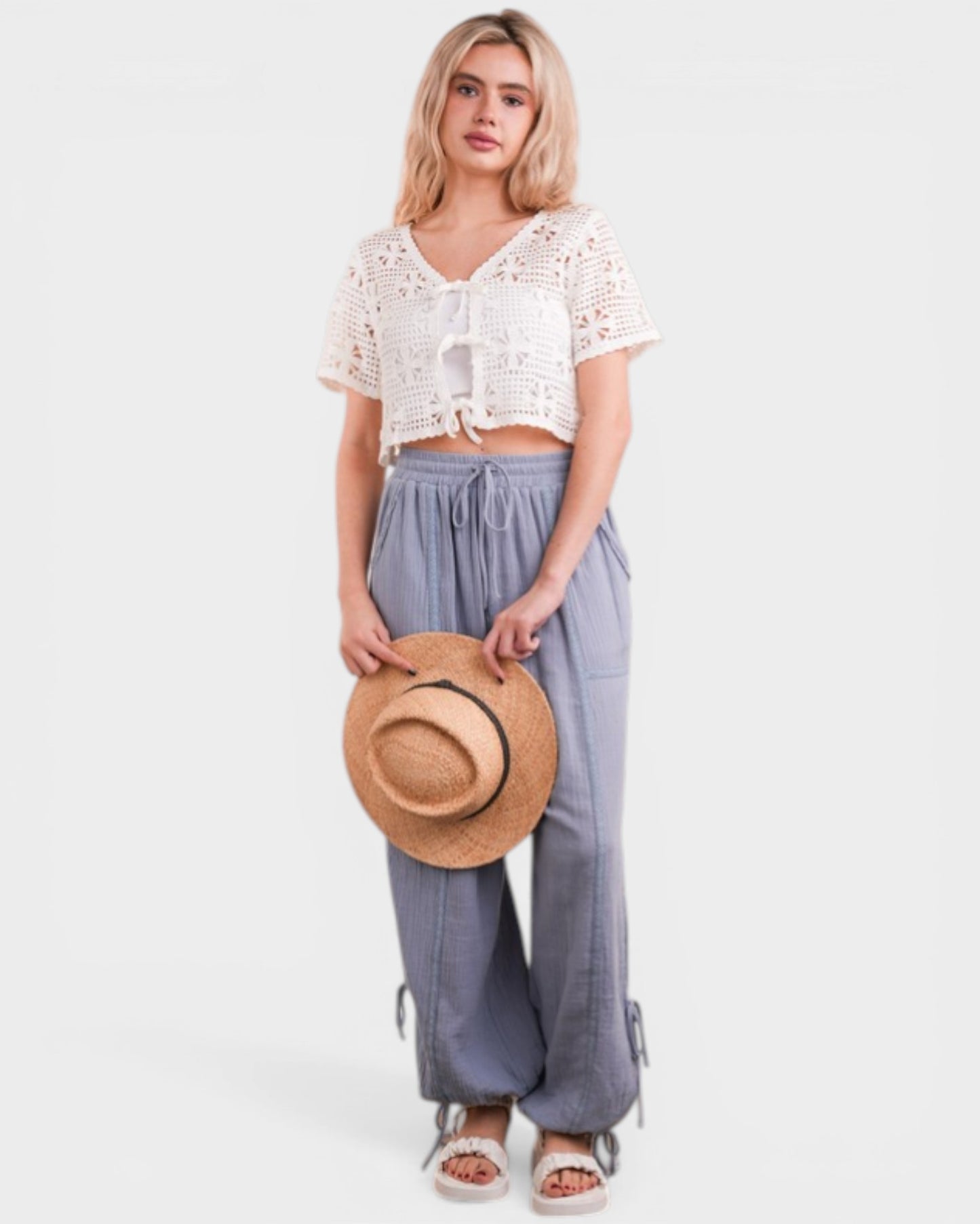 Woman holding a straw hat wearing a white lace top and light blue pants on a white background