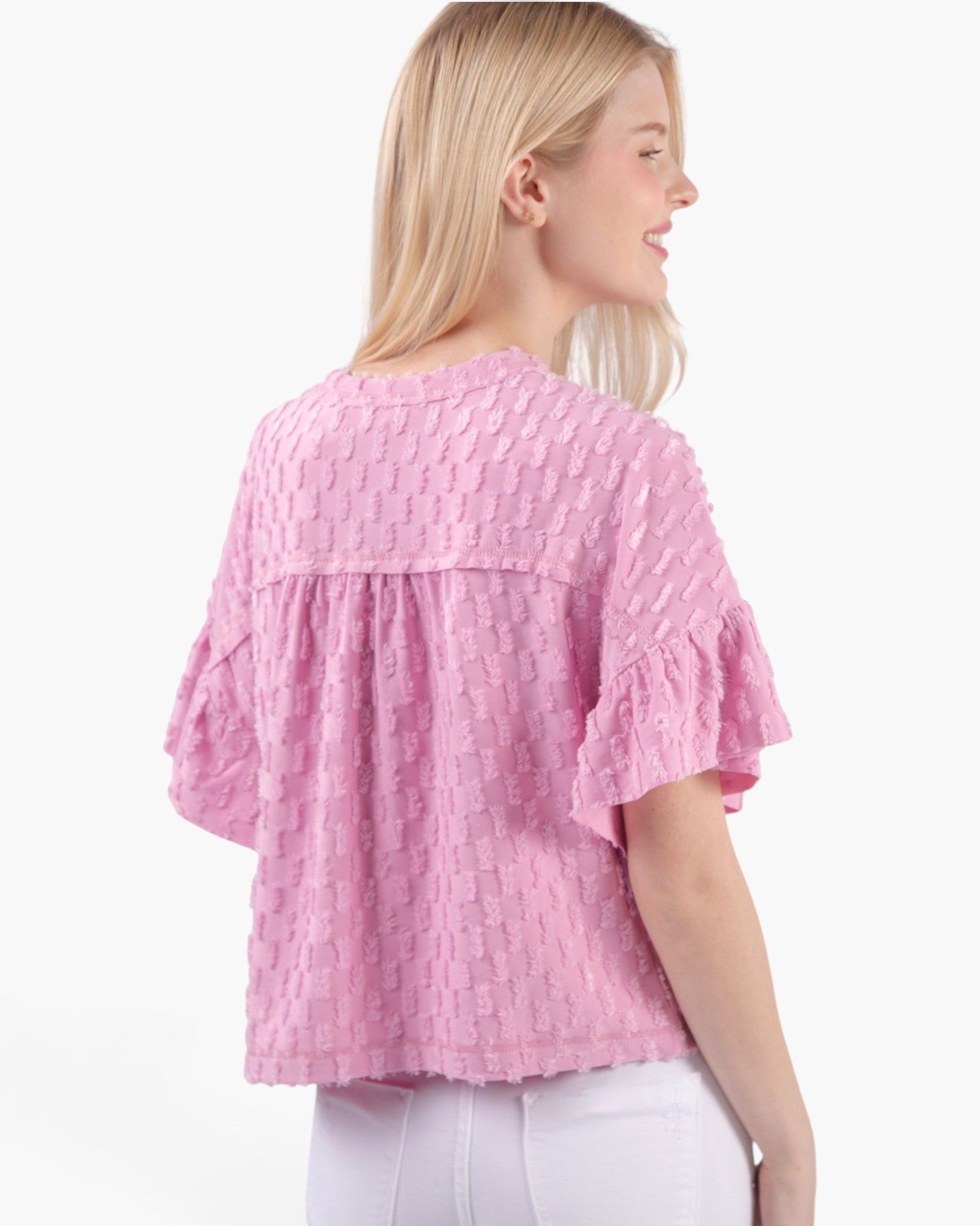 A woman shown from the back wearing a pink textured short-sleeve blouse with ruffled sleeves and white pants, against a plain light gray studio background.