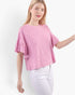 A woman wearing a pink textured short-sleeve top with ruffled details and white pants, photographed against a plain light background.