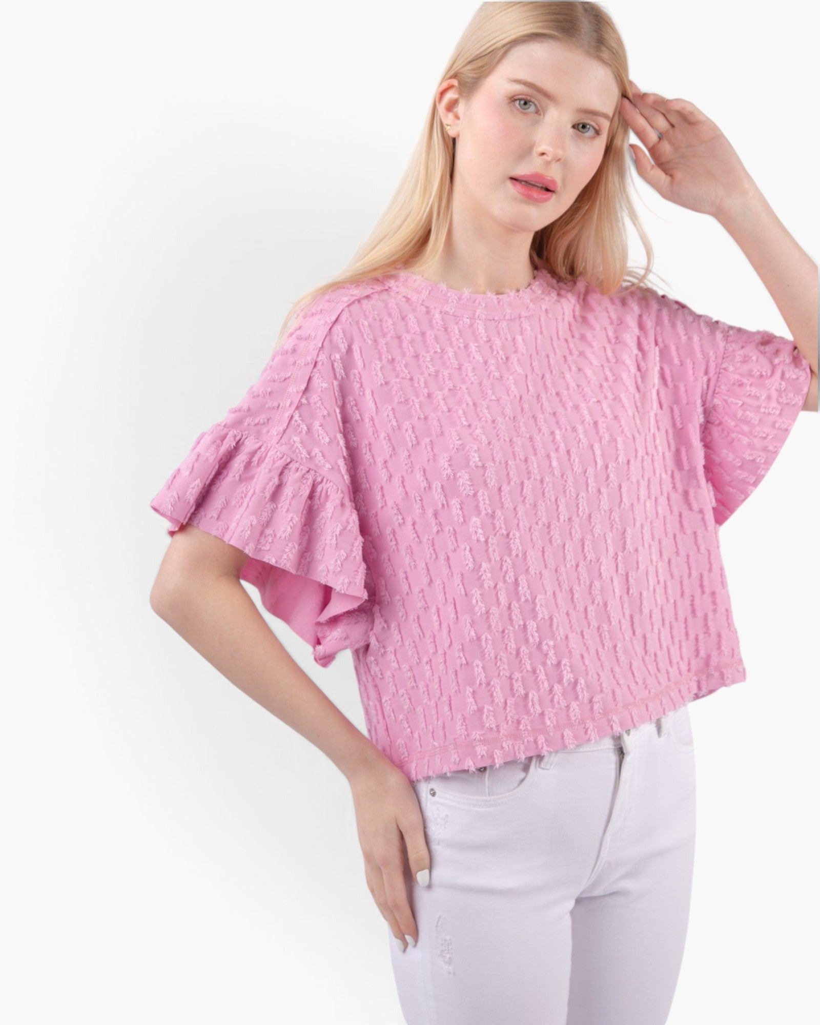 A woman wearing a pink textured short-sleeve top with ruffled flutter sleeves and white pants, photographed against a plain light background.