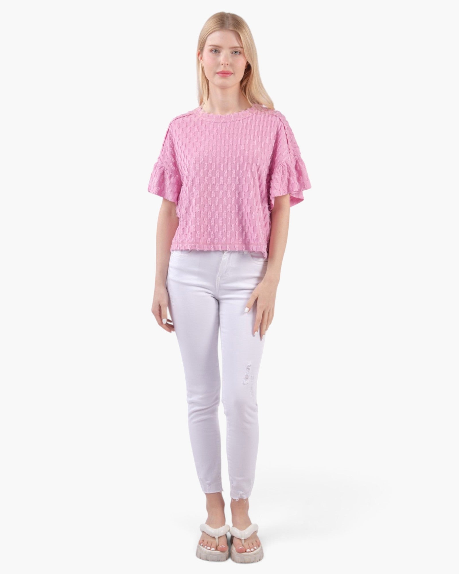 A woman modeling a pink textured short-sleeve top with ruffled sleeves, paired with white skinny jeans and white slide sandals, against a plain white background.