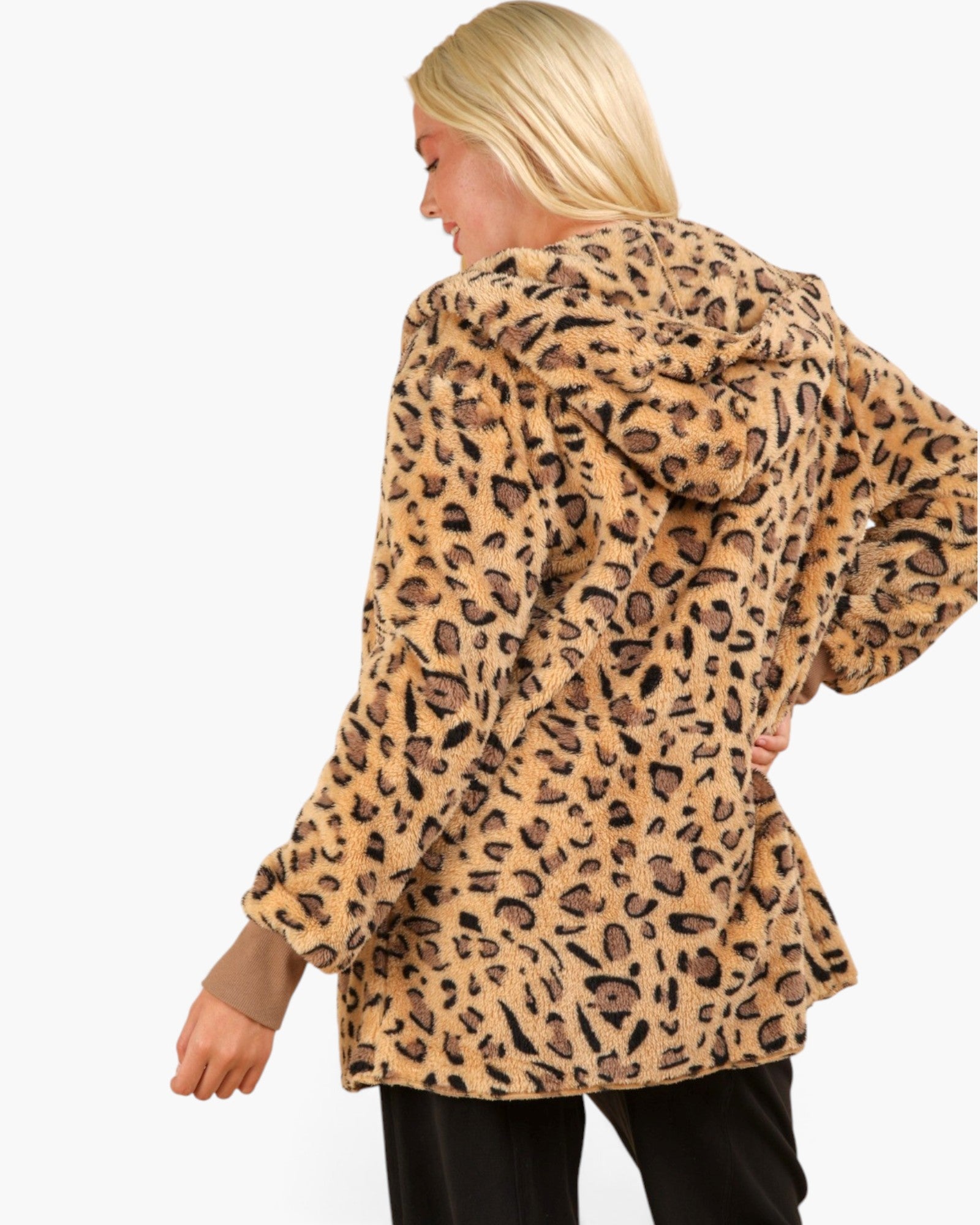 A woman shown from the back wearing a plush leopard-print hooded jacket in tan and brown tones, paired with black pants, against a plain white studio background.