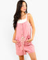 A woman modeling a pink sleeveless romper with a white ribbed tank top underneath, featuring front buttons, a drawstring waist, and front pockets, set against a plain white background.