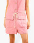A close-up of a model wearing a pink sleeveless romper with a button-down front, large front patch pockets, and a cream-colored rope drawstring at the elastic waistband.