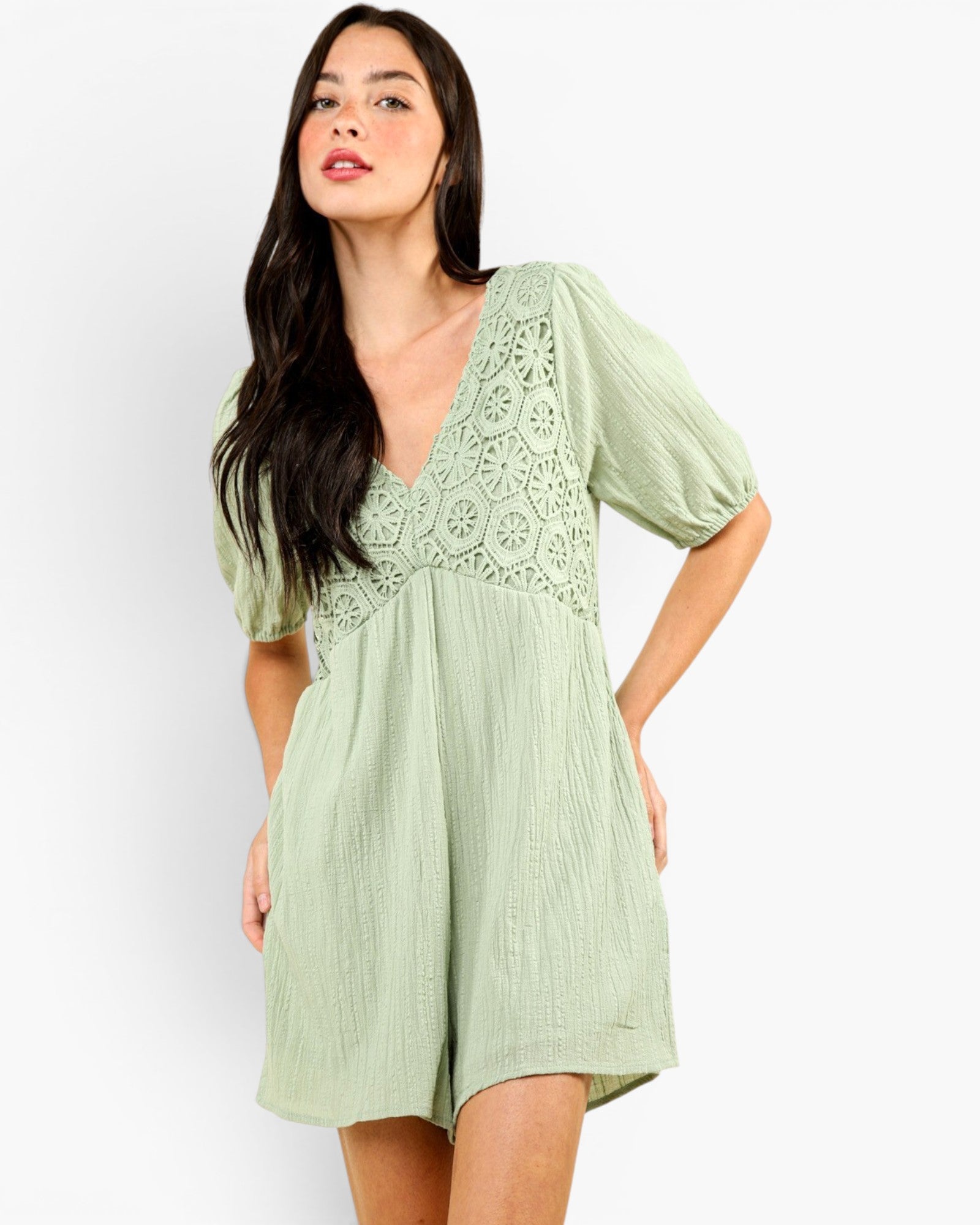 A woman modeling a light sage green crinkle gauze romper with a floral eyelet lace bodice and puff sleeves, set against a plain white studio background.