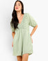 A woman modeling a light sage green crinkle gauze romper with a floral eyelet lace bodice and puff sleeves, set against a plain white studio background.
