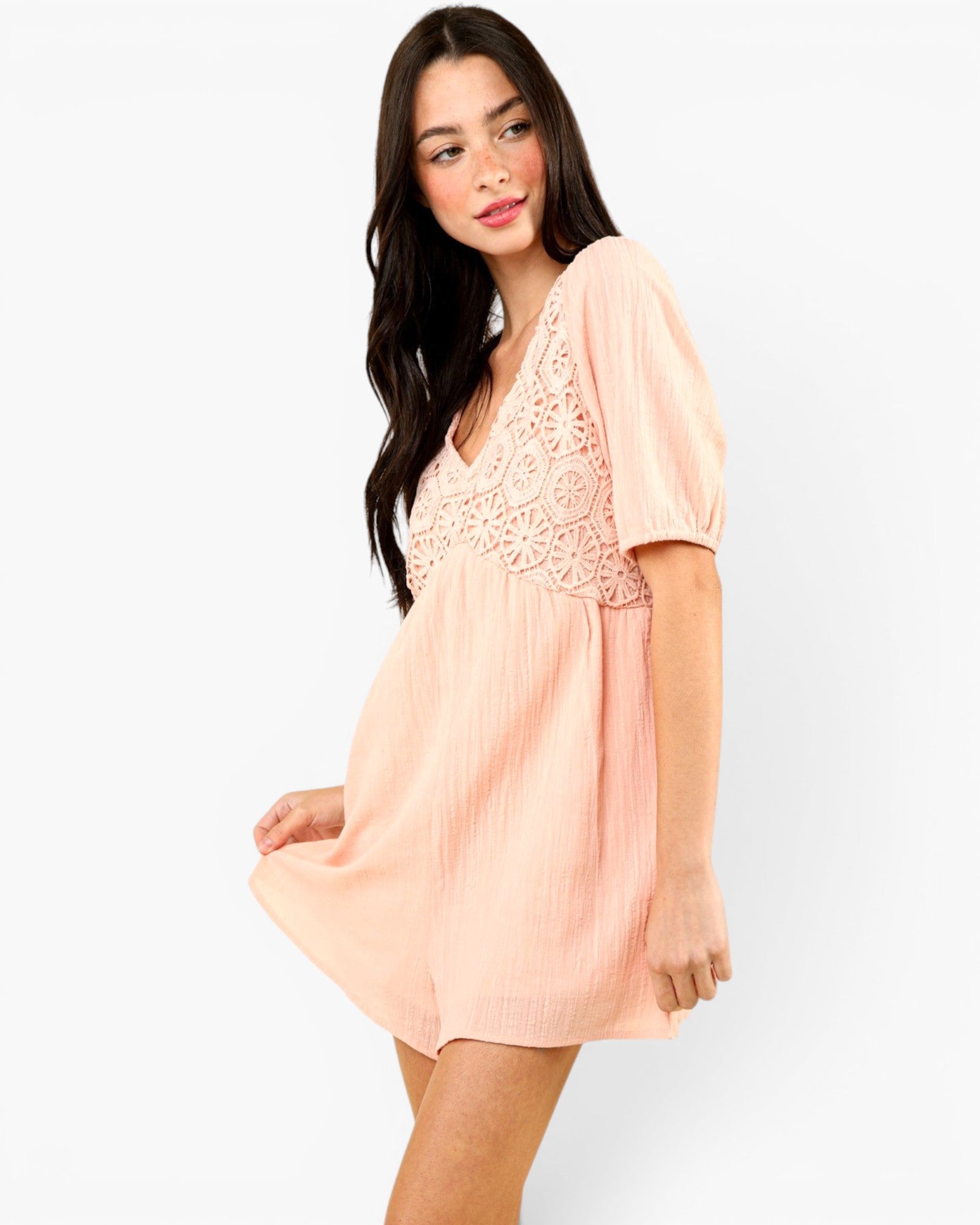 A woman modeling a short peach-pink dress with short puff sleeves, a V-neckline, and a floral lace overlay across the chest, set against a plain white studio background.