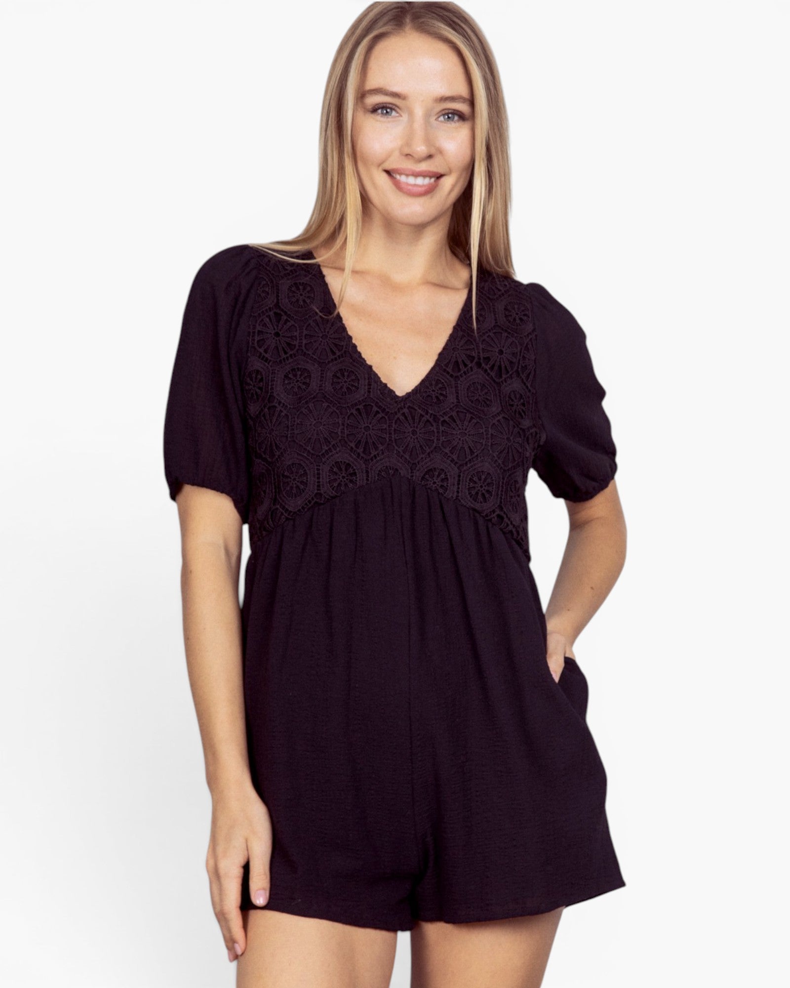 A woman modeling a black short-sleeve romper with a V-neck and intricate lace overlay on the chest, set against a plain white background.
