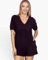 A woman modeling a black short-sleeve romper with a V-neck and intricate lace overlay on the chest, set against a plain white background.