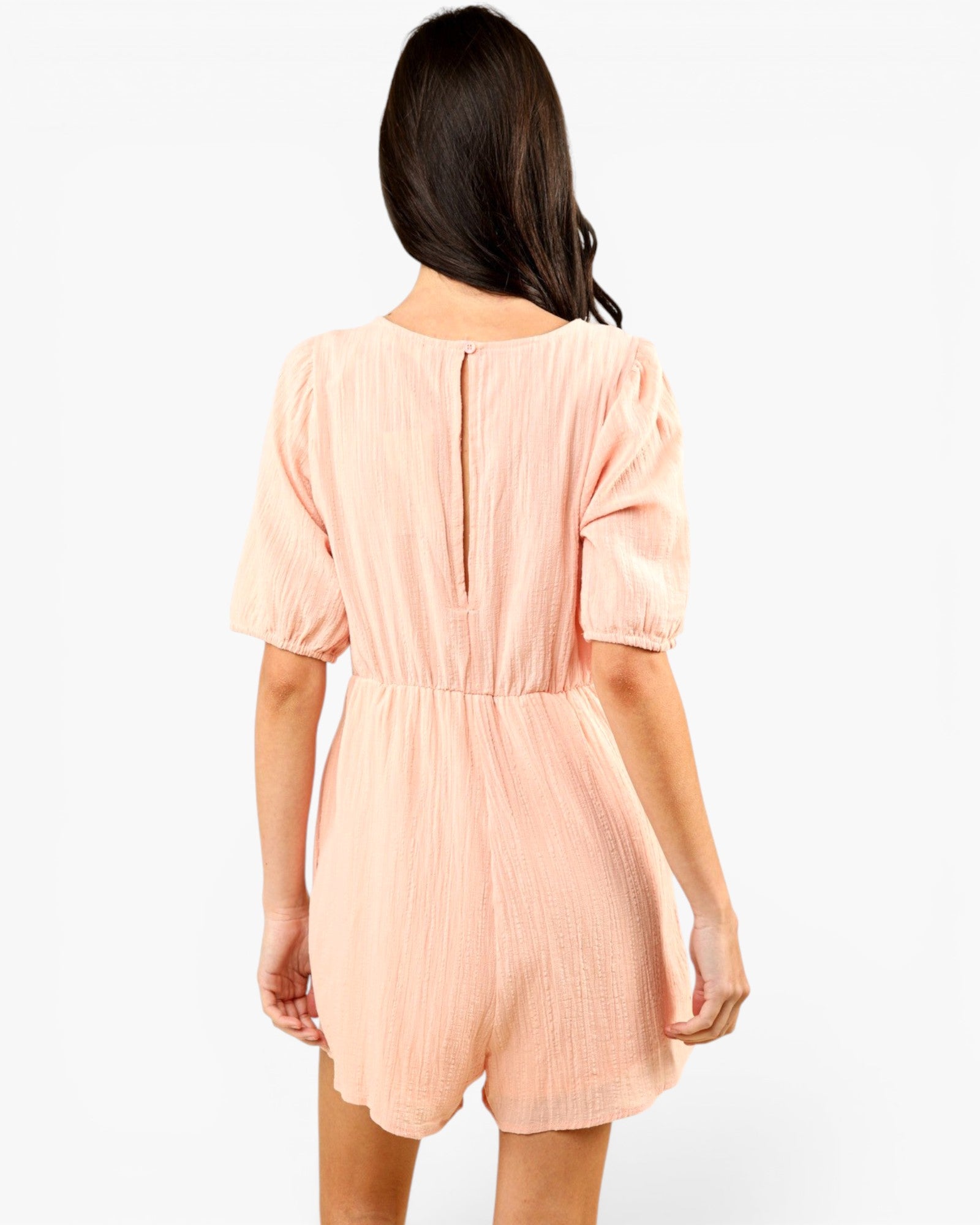 A woman viewed from the back wearing a light peach-pink crinkle-textured romper with short puff sleeves and a gathered waist, set against a plain white studio background.