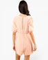 A woman viewed from the back wearing a light peach-pink crinkle-textured romper with short puff sleeves and a gathered waist, set against a plain white studio background.