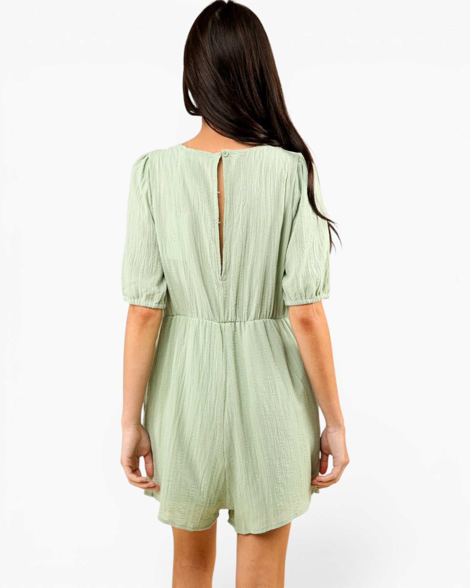 A woman viewed from the back wearing a light sage green crinkle-textured romper with puff sleeves and a back keyhole detail, set against a plain white studio background.